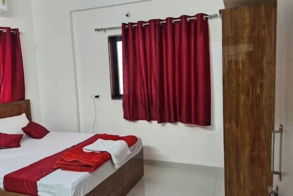 A hotel room with a bed, red curtains, a wooden door, and a small window.
