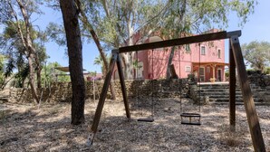 Un conjunto de columpios de madera en un área boscosa, con un muro de piedra y un edificio al fondo.