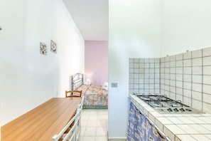 A compact kitchen with a gas stove, a dining table with chairs, and a bed with a colorful bedspread in the background.