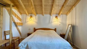 Een slaapkamer met een houten plafond, een bed met een witte spreideken, twee wandlampen, een houten stoel en een klein tafeltje.