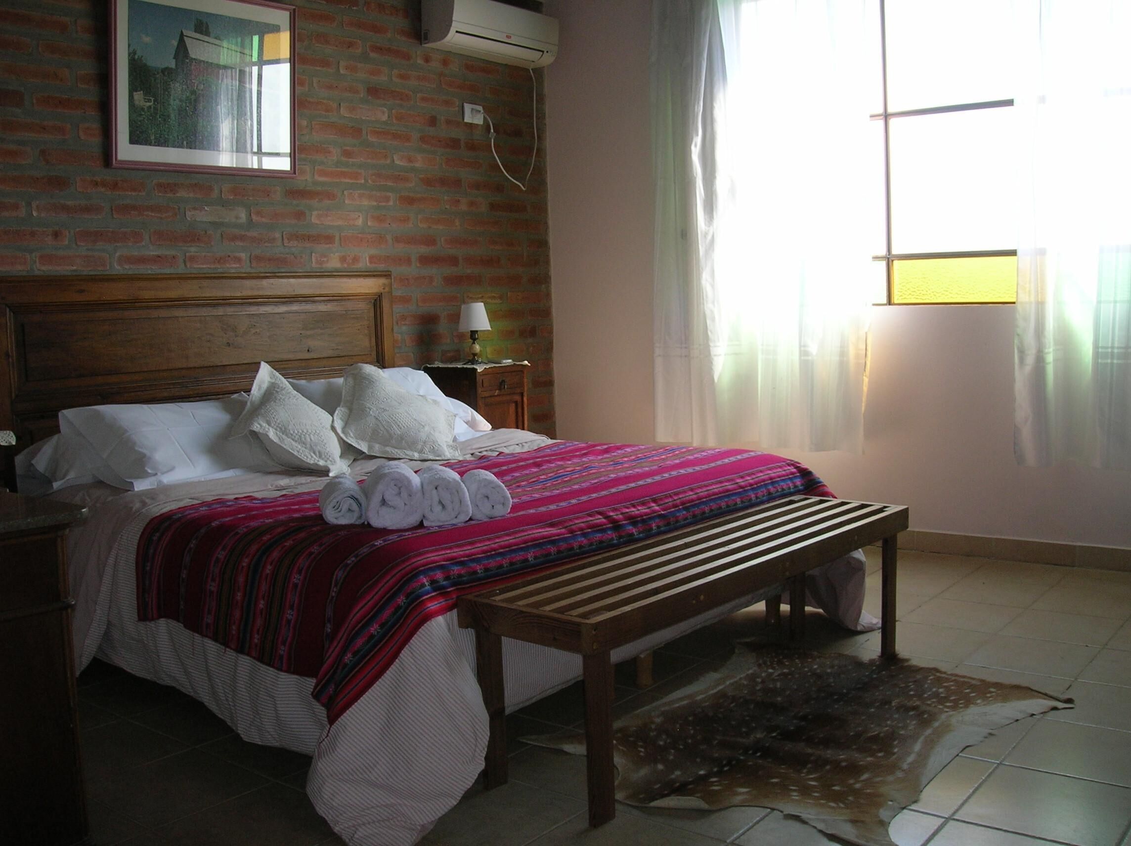 A bed with rolled towels, a wooden bench, a brick wall, a framed picture, and a window with curtains.