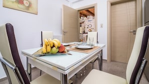 A dining area with a table set for two, featuring a fruit platter, a glass, and a plate. The room has a white wall with a painting, a wooden door, and a view into another room with a brick wall and a clock.