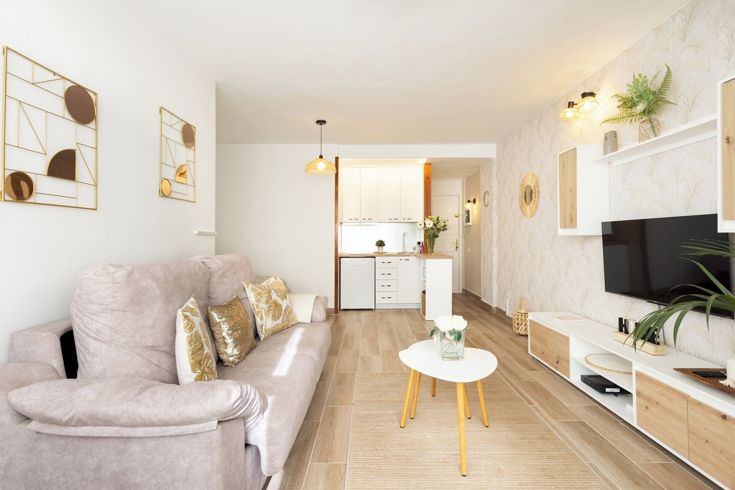 A modern living room with a sofa, a coffee table, and a TV. There is a kitchen in the background with white cabinets and a countertop.