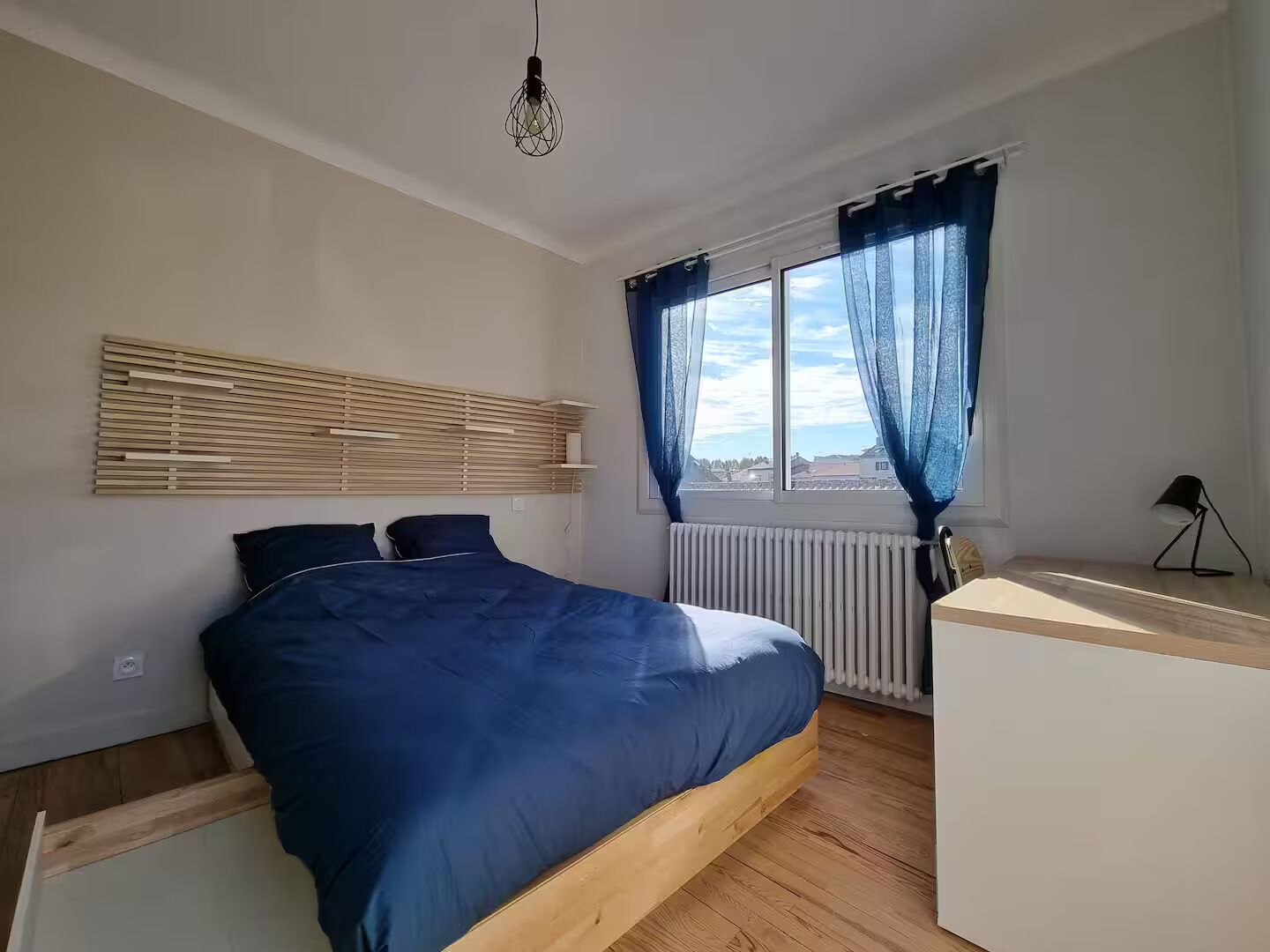 Ein Schlafzimmer mit Bett, Holz-Kopfteil, Fenster mit Vorhängen, Heizkörper und Schreibtisch mit Lampe.