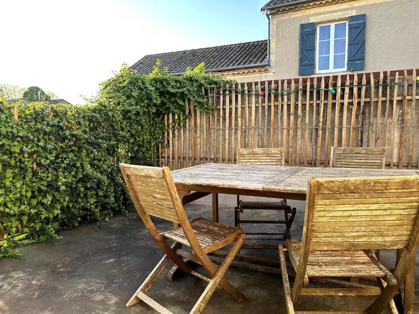 A wooden table and chairs set outdoors with a wooden fence and greenery in the background.