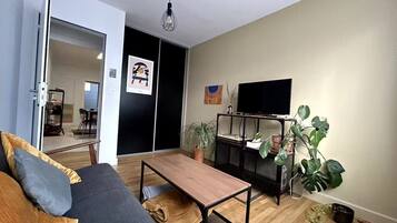 A modern living room with a sofa, coffee table, and a TV on a stand. There are plants and a door leading to another room.
