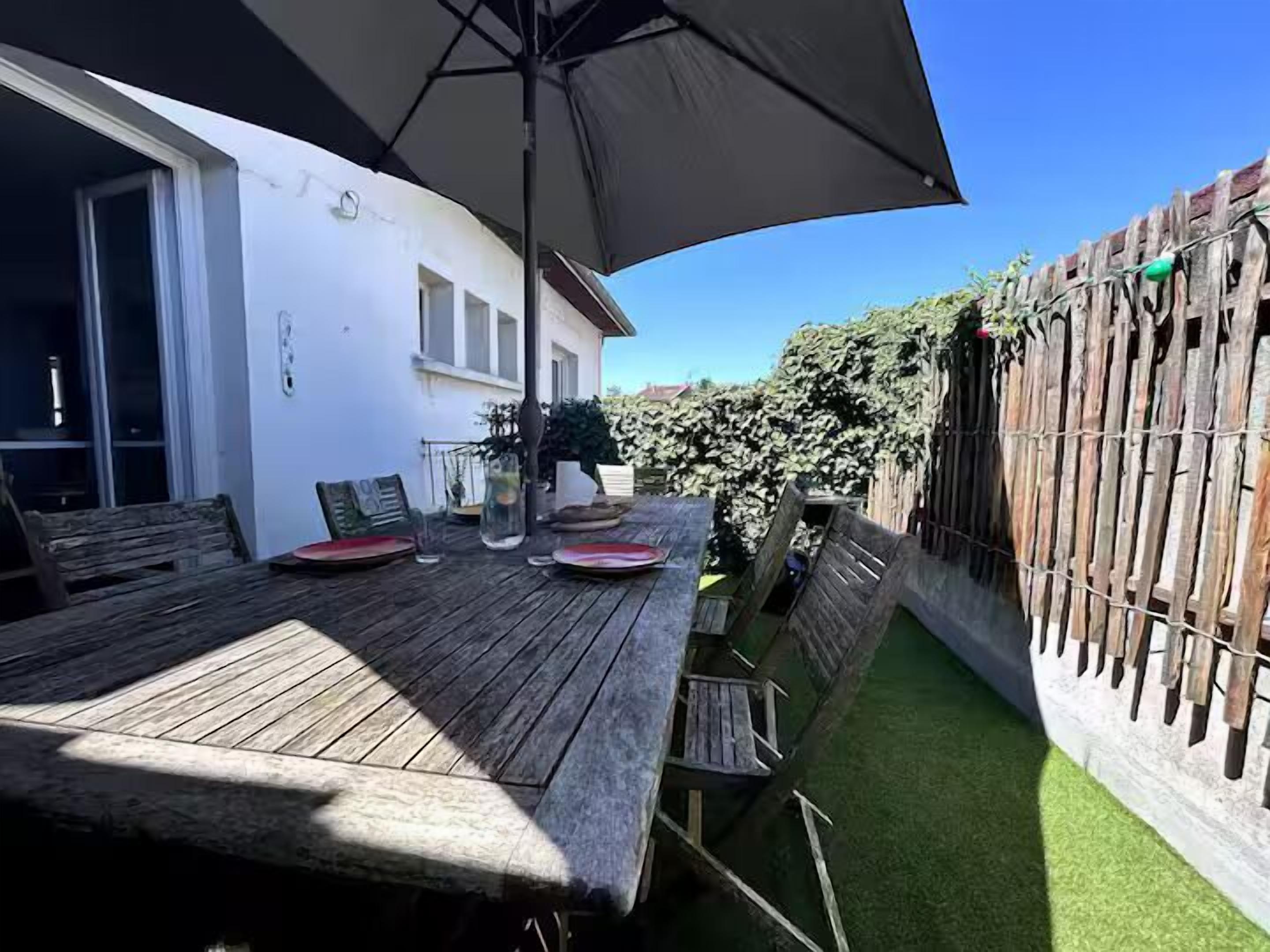 Una terraza de madera con una mesa, sillas y un paraguas grande, y un edificio blanco al fondo.