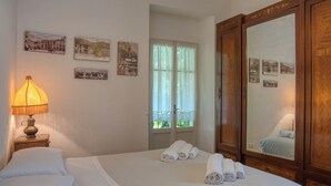 Een hotelkamer met een bed, nachtkastje met lamp, raam met gordijnen en spiegel met foto's aan de muur.
