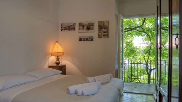 Een slaapkamer met een bed, een nachtkastje met een lamp, en een balkon met uitzicht op groen.