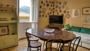 Een eethoek met een tafel, stoelen en een televisie. Er staan planken met servies en een raam met uitzicht op de bergen.