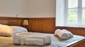 Una cama bien hecha con toallas enrolladas, cabecera de madera, ventana con vistas a la vegetación y una lámpara en la mesita de noche.