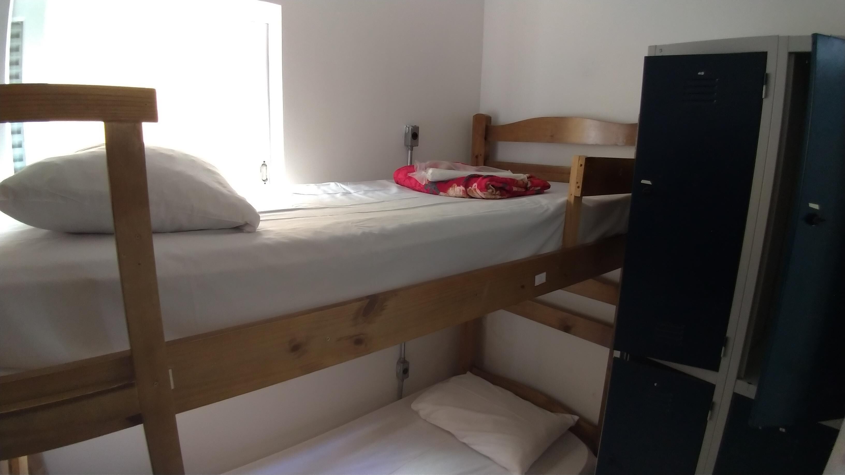 Una cama litera con colchón y almohadas blancas, una colcha roja y un cabecero de madera. A la derecha, un escritorio pequeño con una silla y un armario negro.