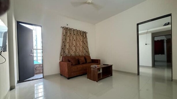 A spacious room with a large window, a brown sofa, a wooden coffee table, and a ceiling fan. - 1BHK Comfy Stay 202 (Pune)