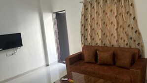 A living room with a brown sofa, a flat-screen TV mounted on the wall, and a patterned curtain. - 1BHK Comfy Stay 202 (Pune)