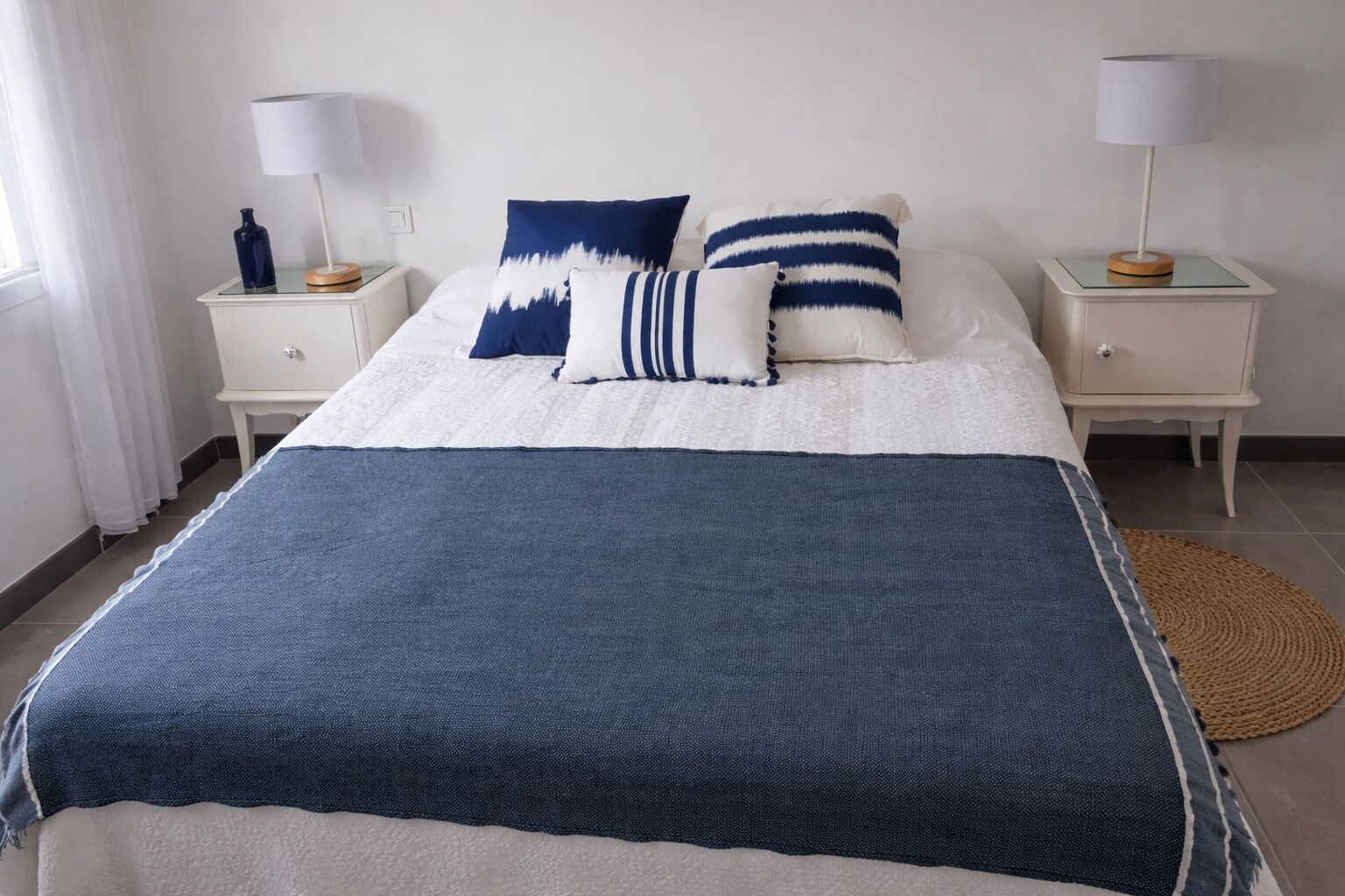 A bed with a blue and white bedspread, two bedside tables with lamps, and a woven rug on the floor.
