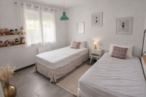 A bedroom with two beds, a bedside table with a lamp, a shelf with plush toys, and a window with curtains.
