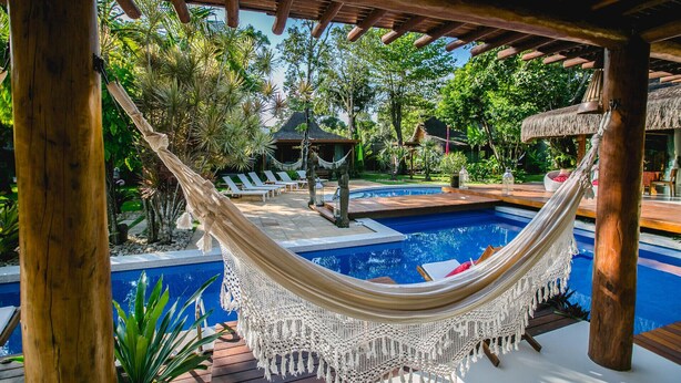 A hammock under a wooden pergola over a swimming pool surrounded by lounge chairs and tropical plants. - Legian Villa Hotel (Porto Seguro)