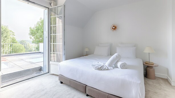 A bedroom with a large bed, a sliding glass door to a balcony, and a small bedside table with a lamp.
