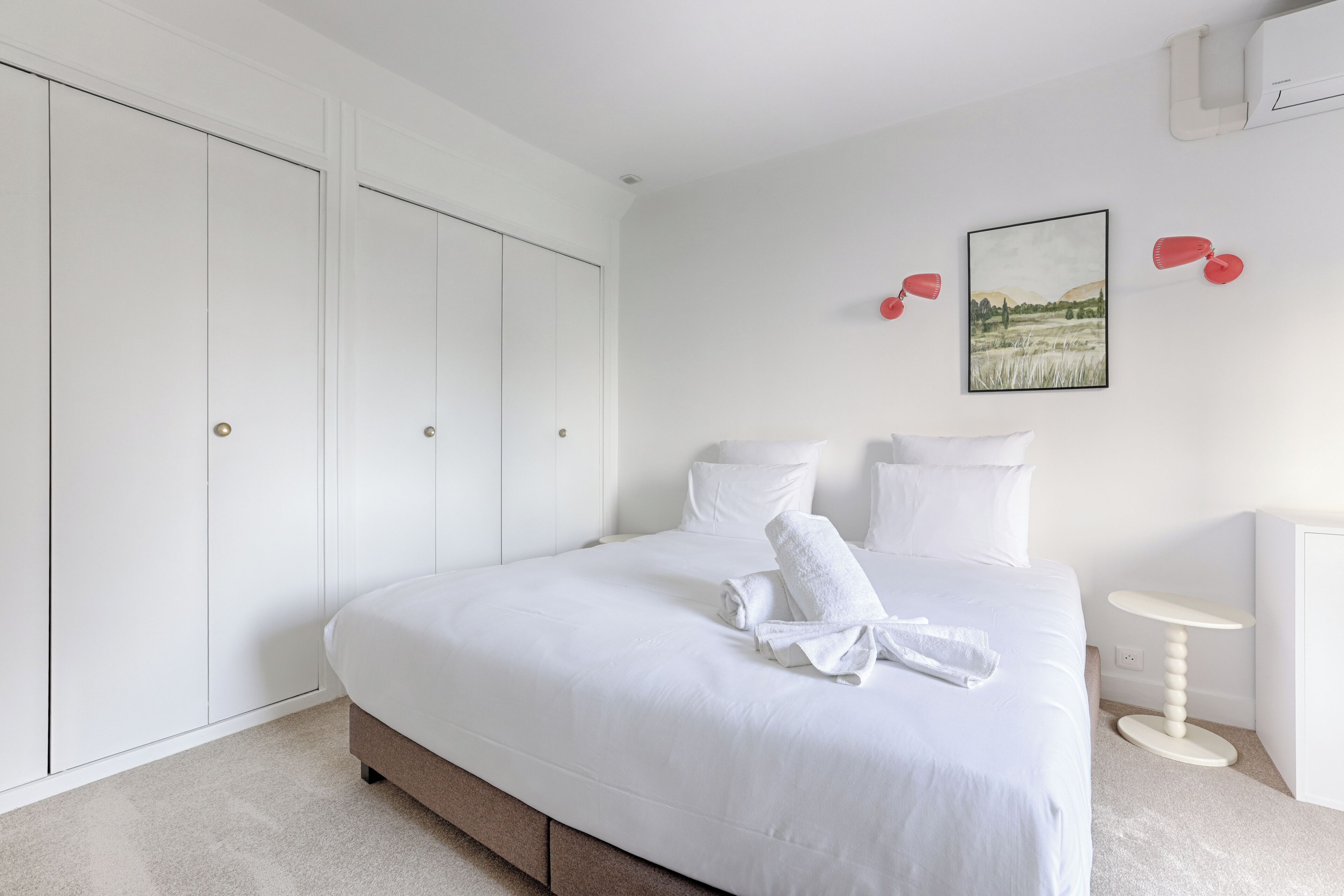Een keurig opgemaakt slaapkamer met een groot bed, een garderobe, een klein tafeltje en een wandlamp.