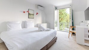 A modern bedroom with a large bed, a desk, and a balcony with a view of trees.
