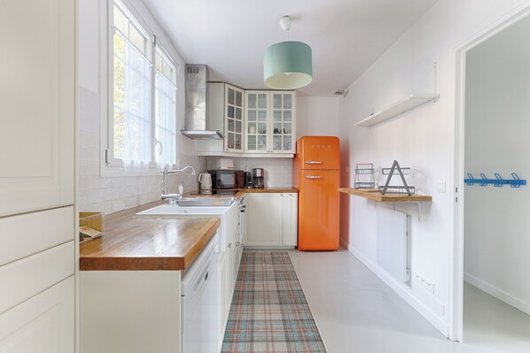 A kitchen with white cabinets, a wooden countertop, a plaid floor mat, a teal pendant light, and a vintage orange refrigerator.