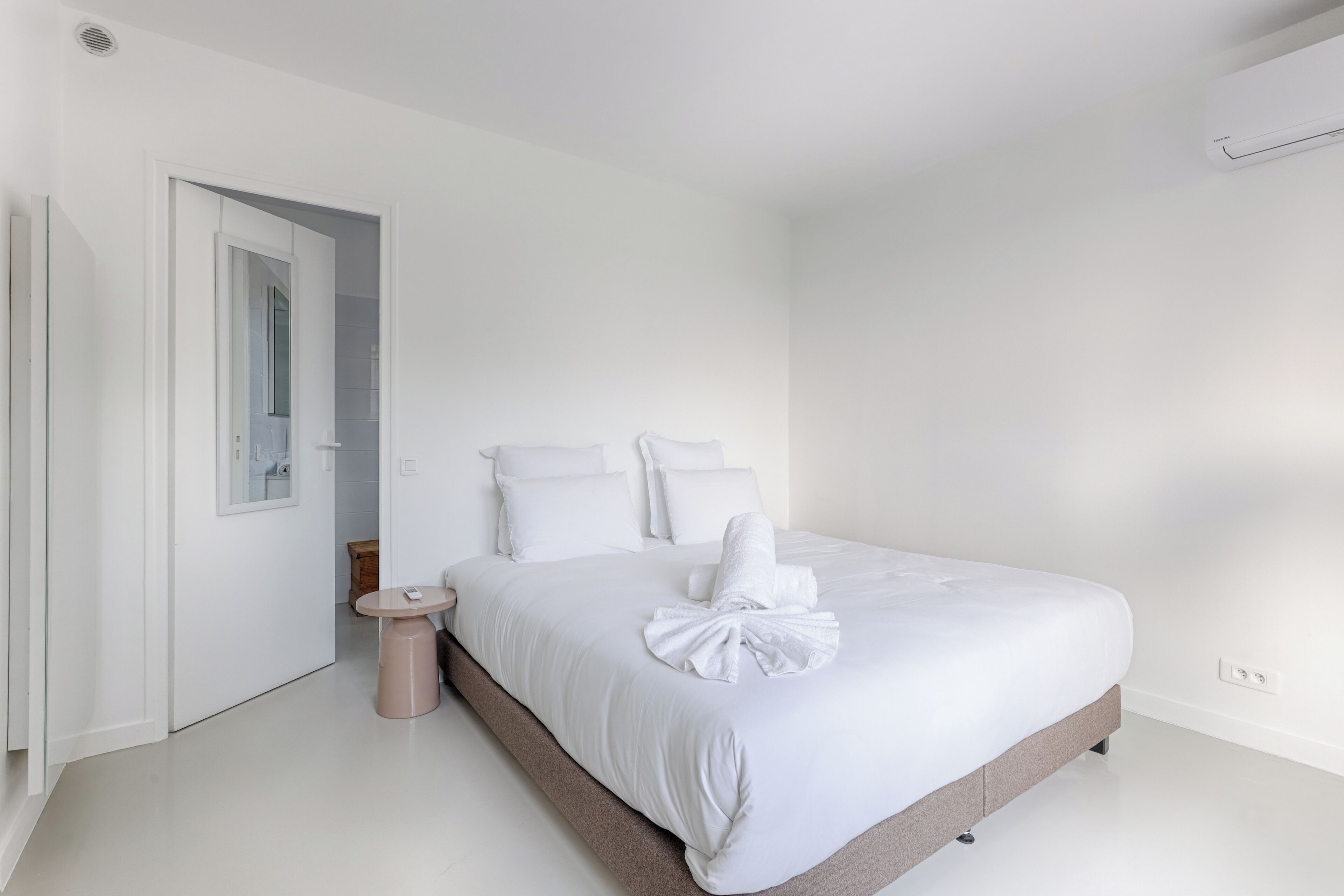 A minimalist hotel room with a large bed, a bedside table, and a small stool.