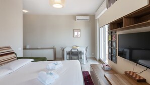 Quarto de hotel com cama, escrivaninha, cadeira, televisão e uma janela grande.