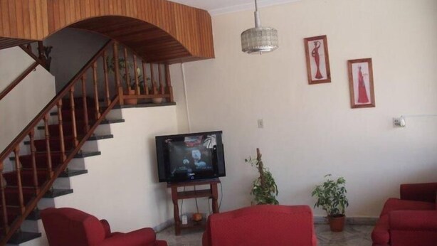 A living room with a wooden staircase, a flat-screen TV on a stand, two red sofas, and two potted plants. - Hotel Compostela (Necochea)