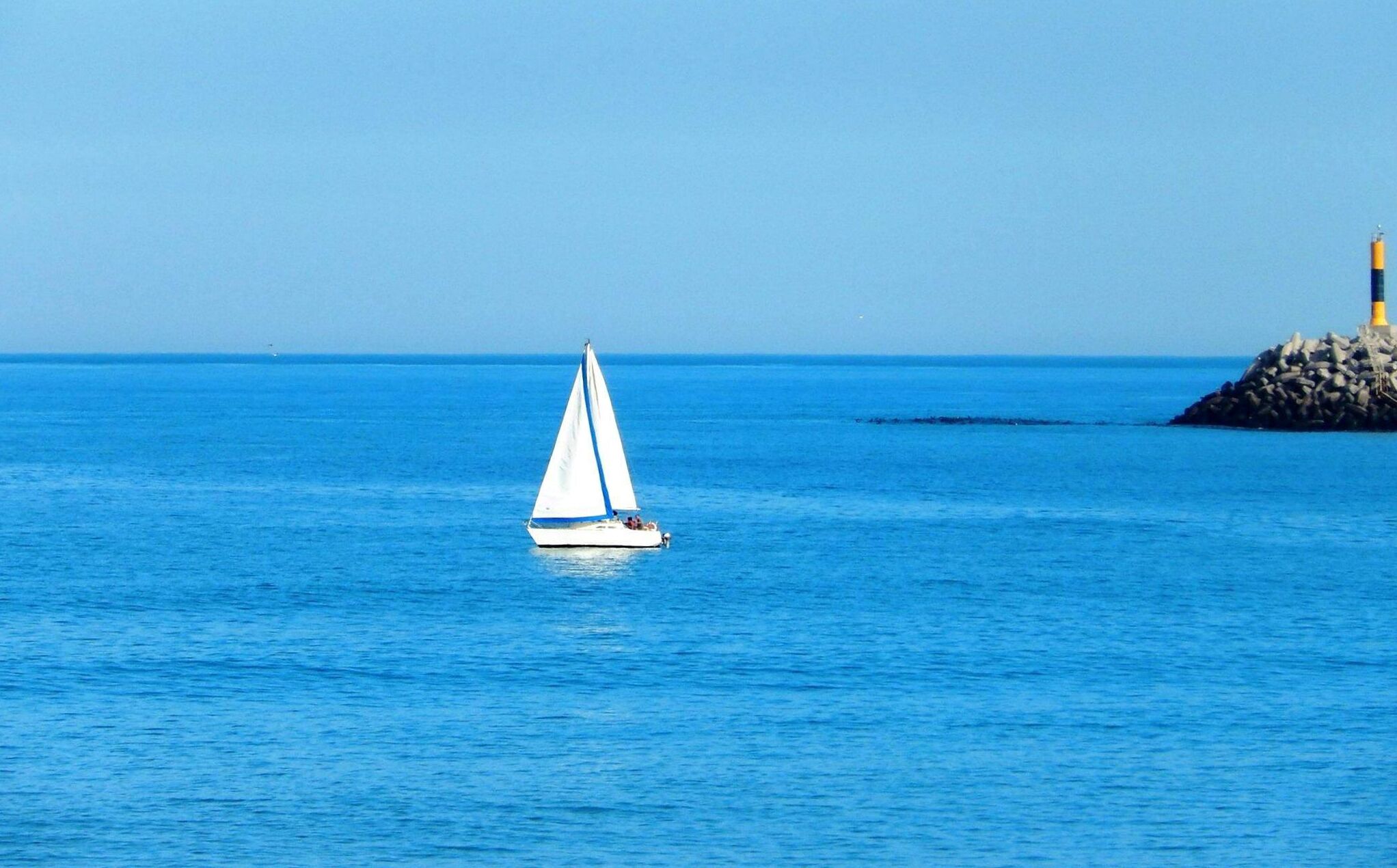 Una barca a vela su acque blu calme, con un promontorio roccioso e un faro in lontananza.