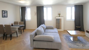 A modern living room with a grey sofa, a dining table with chairs, and a small cabinet. - Margot (Vienna)