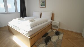 A modern bedroom with a wooden bed, white bedding, a small bedside table, and a view of the outdoors through a window. - Margot (Vienna)