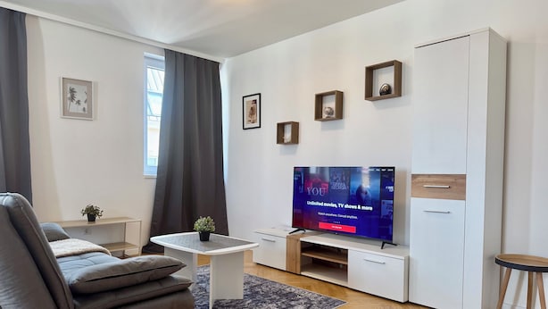 A modern living room with a flat-screen TV, grey sofa, small side table, and a white cabinet. - Margot (Vienna)