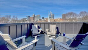 A rooftop deck with two Adirondack chairs and a small table, overlooking a city skyline.