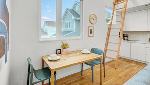 A modern kitchen with white cabinets, a wooden table, and a blue chair. A ladder is leaning against the wall, and a clock is visible on the wall.