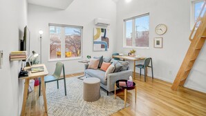 A modern living room with a loft-style ceiling, a grey sofa with multiple cushions, a wooden dining table, and a large window providing a view of the outside.