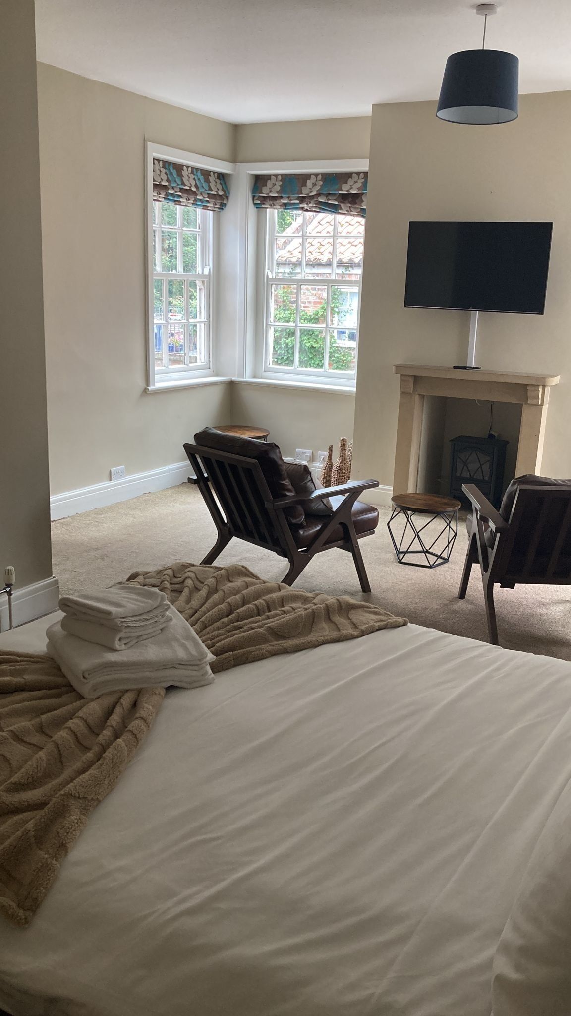 A bedroom with a bed, two chairs, a TV, a fireplace, and windows with curtains.