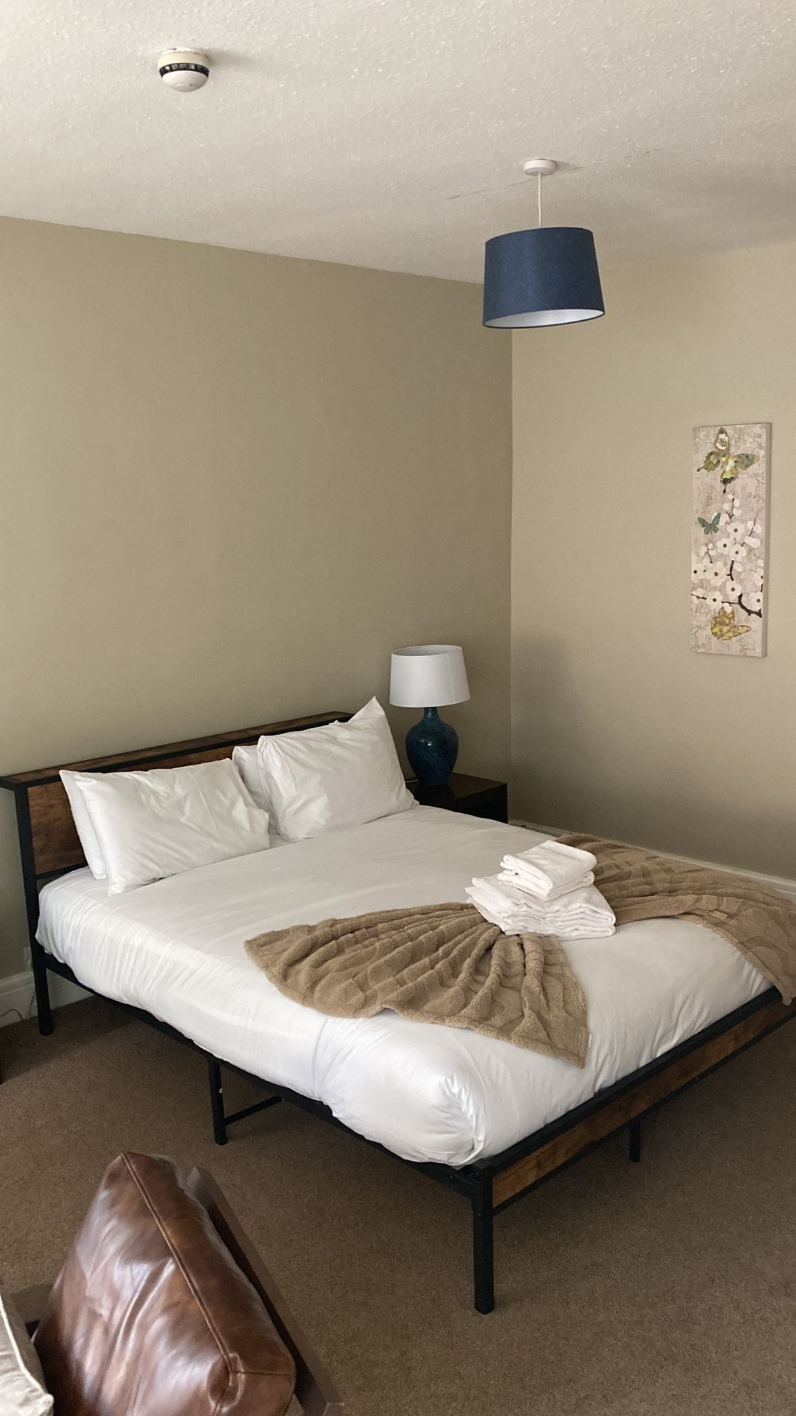 A neatly made bed with white linens and a brown throw blanket, a bedside lamp, a wall-mounted artwork, and a ceiling-mounted smoke detector.