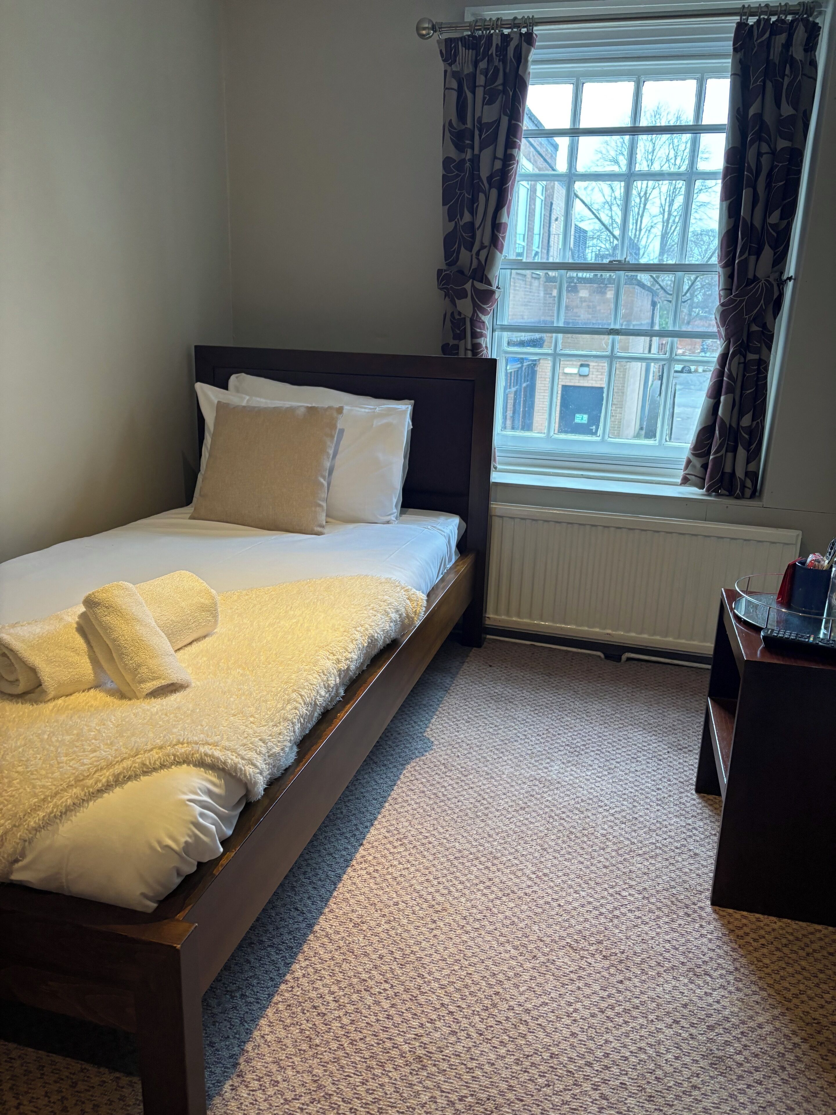 A neatly made bed with white linens and a fluffy white blanket, a wooden headboard, a window with patterned curtains, and a radiator below.