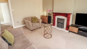 A living room with a beige sofa, a small round table, a fireplace, and a television. - 3 Hawk Place (Whitehaven)