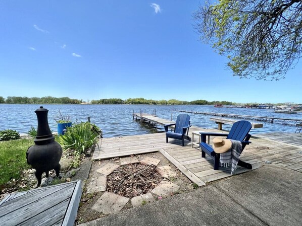 Une jetée en bois, agrémentée de chaises et d’un foyer, d’où l’on peut admirer un lac paisible avec des bateaux au loin.