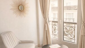 A room with a white chair, a round table with a book and a cup, a sunburst mirror, and a view of balconies through a window with curtains. - 1565 Pétion (Paris)