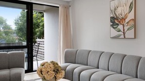 Un salon moderne avec un canapé gris, une petite table sur laquelle est posé un vase de fleurs et une grande fenêtre donnant sur un paysage verdoyant.