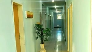 A long, narrow hallway with light blue walls and a tiled floor. There are doors on both sides, a potted plant in the center, and a ceiling light. - DC Hostel (Florianópolis)
