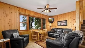 A wooden-paneled room with a dining area, two black leather sofas, a ceiling fan, and a television on the wall. - Cabin 1 at Heath's Resort (Pine River)