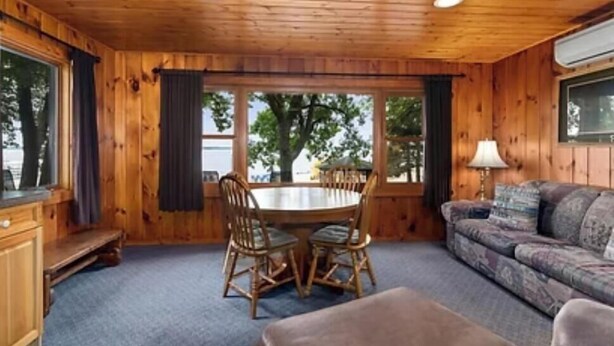 A wooden cabin interior with a dining table, two chairs, a sofa, and a window with a view of trees.