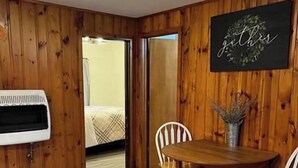 A room with wooden walls, a bed with a plaid blanket, a white air conditioner, a wooden dining table with chairs, and a sign with the word "gether". - Cabin 6 at Heath's Resort (Pine River)