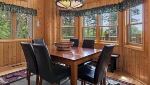 Interior de una cabaña de madera con una mesa de comedor, cuatro sillas, una araña y ventanas amplias.