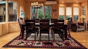 A wooden interior with a dining area featuring a chandelier, a rug with animal prints, and large windows offering a view of the outdoors.