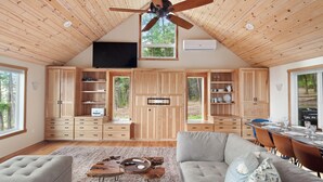 A spacious living area with a high ceiling, wooden ceiling, and a large window. There is a flat-screen TV mounted on the wall, a dining table with chairs, and a sofa with cushions.
