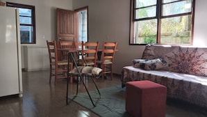 A room with a dining table and chairs, a sofa with patterned cushions, a red ottoman, and a view of greenery through the windows.